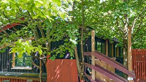 A wooden play structure with slides and a fenced area, surrounded by trees and a building with a window. - Gorgeous apartment in åsljunga (Åsljunga)