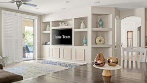 A living room with a flat-screen TV, white shelving, and a ceiling fan.