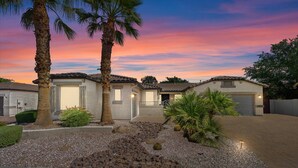 A residential area with two houses, a driveway, and a well-maintained garden with palm trees and shrubs.