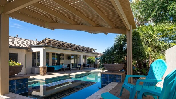 A covered outdoor swimming pool area with lounge chairs, a patio, and a building with a tiled roof in the background.