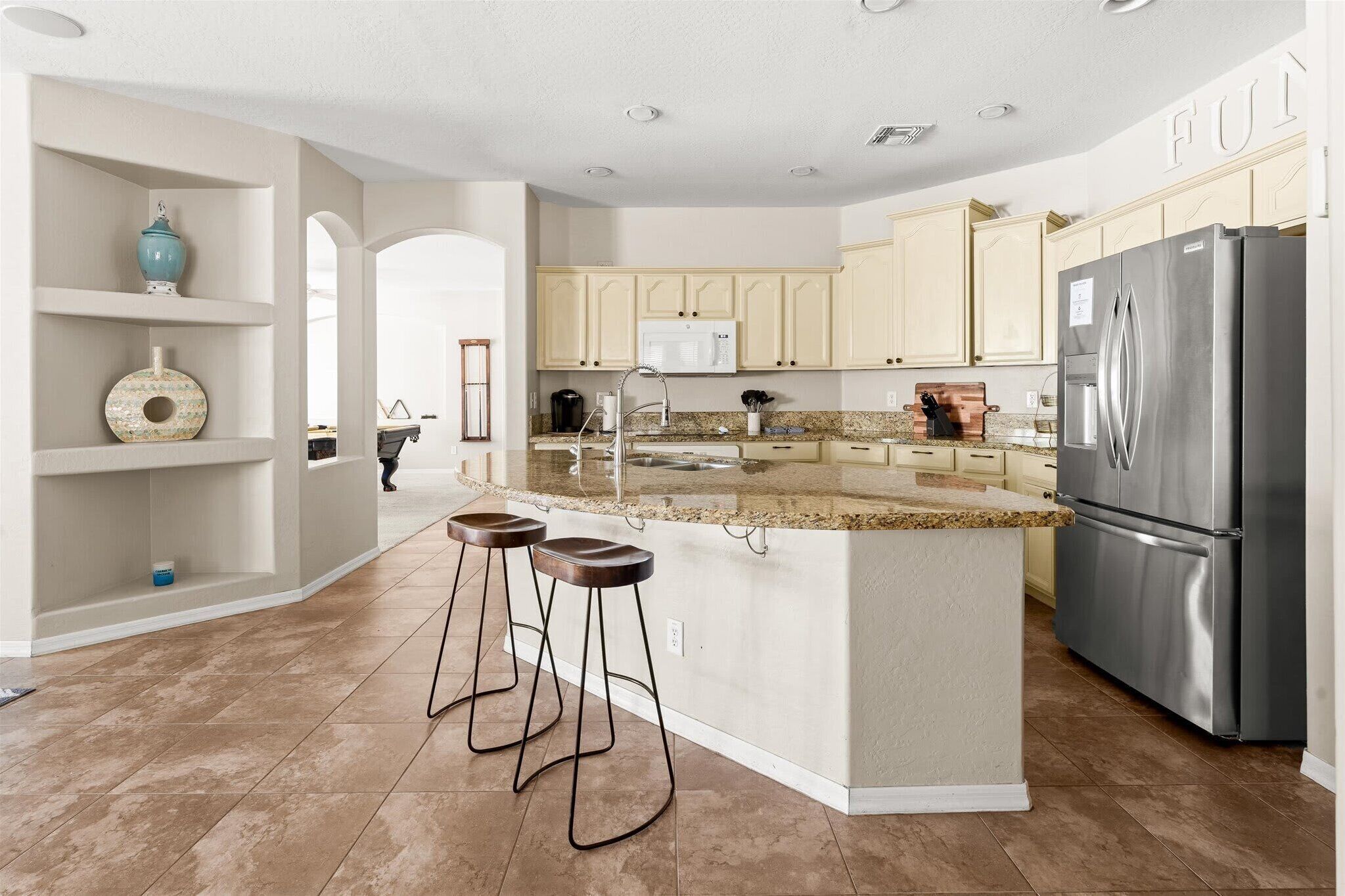 A modern kitchen with a central island, bar stools, a stainless steel refrigerator, and light-colored cabinetry.
