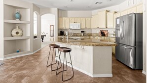 A modern kitchen with a central island, bar stools, a stainless steel refrigerator, and light-colored cabinetry.