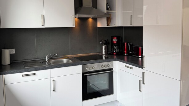 A modern kitchen with white cabinets, a stainless steel range hood, a built-in oven, and a black countertop. - Vacation apartment Klaus (Clausen)