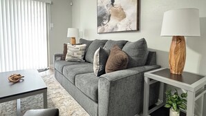 Een moderne woonkamer met een grijze bank, een bijzettafeltje, een lamp en een schilderij aan de muur.
