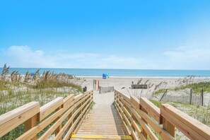 Une promenade en bois mène à une plage où l’on aperçoit un parasol bleu et une clôture au loin.