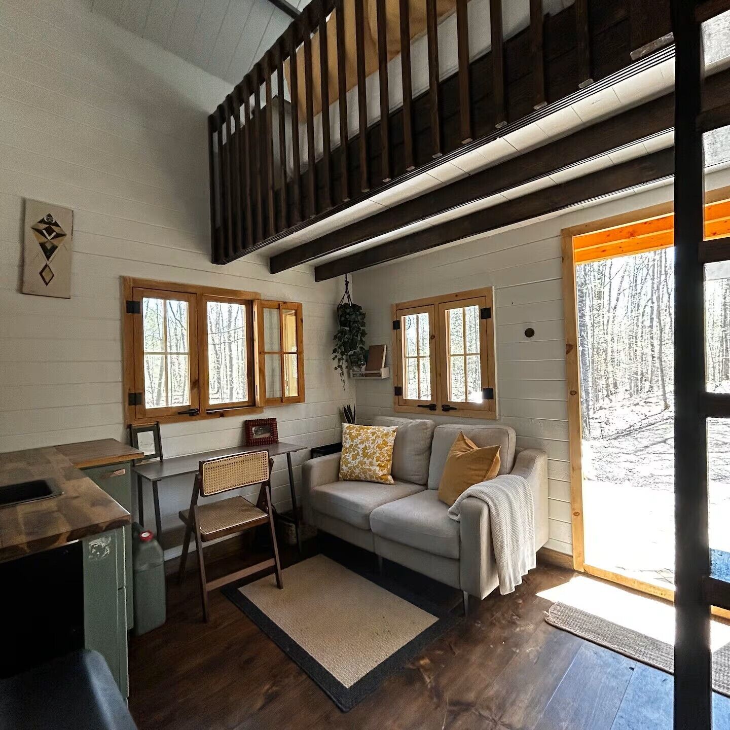 A cozy interior with a wooden staircase, a sofa with cushions, a dining table, and a view of a snowy landscape through a large window.