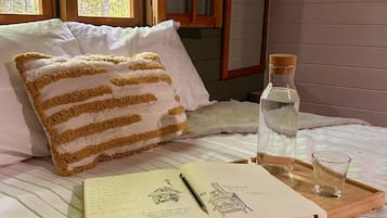 Een slaapkamer met een bed, een raam, een kussen, een boek, een pen, een glas en een fles.