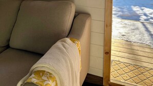 Una ventana con marco de madera que da a un paisaje nevado, un sofá color beige con una manta tejida blanca, y una terraza de madera afuera.