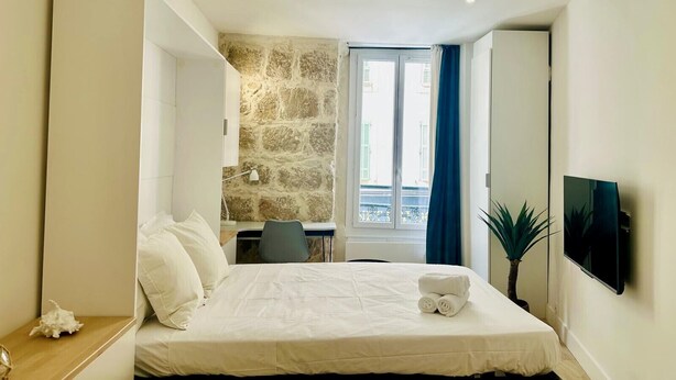 A modern hotel room with a large bed, a desk, a stone wall, a window with curtains, and a TV on the wall. - Cocon Lamartine (Niza)