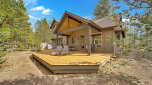 Une maison moderne dotée d’une terrasse en bois, d’un espace salon extérieur et d’une grande fenêtre donnant sur une zone boisée.
