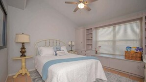 A bedroom with a bed, bedside tables, a lamp, a ceiling fan, a window with blinds, and a built-in shelf unit.