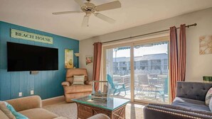 A living room with a flat-screen TV, a ceiling fan, a sign reading "BEACH HOUSE", and a view of a building through large glass doors.