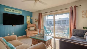 A living room with a flat-screen TV, a ceiling fan, a sign reading "BEACH HOUSE", and a view of a building through large glass doors.
