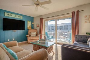 Ein Wohnzimmer mit Flachbildfernseher, Deckenventilator, einem Schild mit der Aufschrift 'BEACH HOUSE' und Blick auf ein Gebäude durch große Glaswände.
