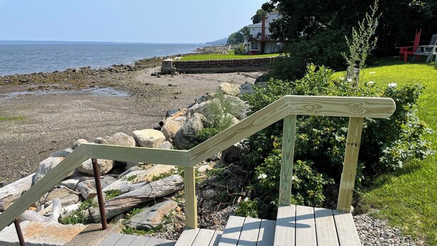 En trätrappa leder ner till ett område vid stranden, med utsikt över havet och ett hus i bakgrunden.