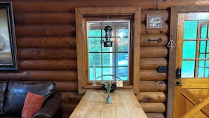 Interior de una cabaña de madera con una mesa de madera, un sofá de cuero negro y una ventana con un farol.