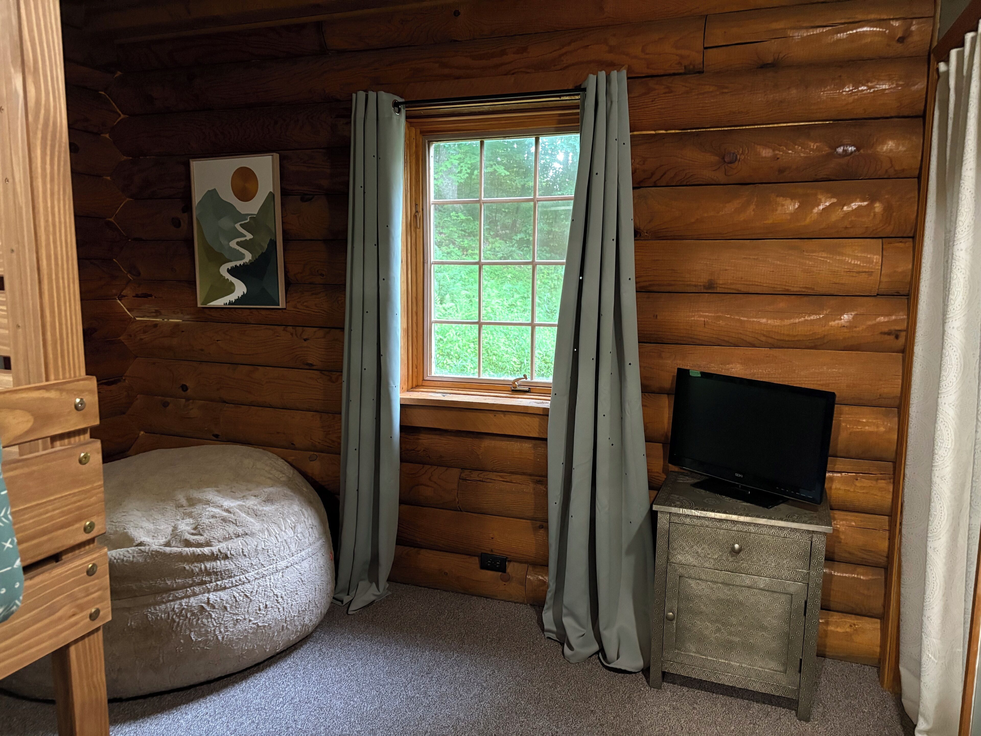 A cozy cabin room with a large window, a bean bag chair, a small wooden cabinet, and a framed picture on the wall.