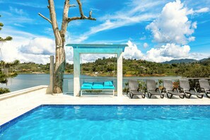 Una piscina con bancos y sillas, una pérgola y vistas a un lago y colinas.