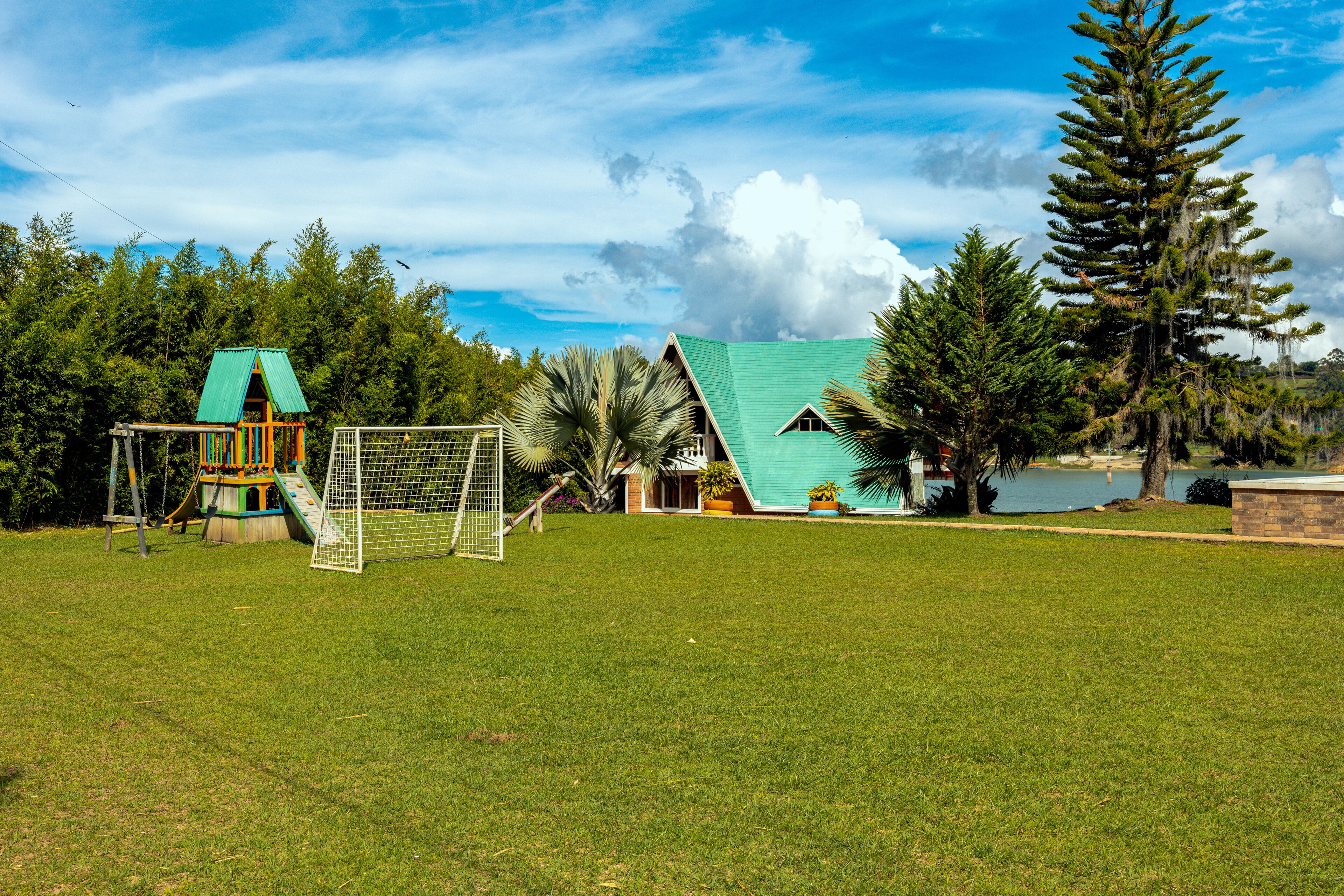 A grassy field with a playground, a small building with a green roof, and a large tree.