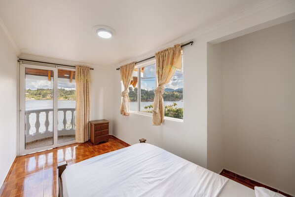 Un dormitorio con cama, cómoda de madera, mesita de noche pequeña, ventana con cortinas y balcón con vista al paisaje.