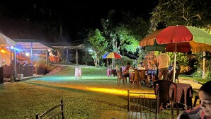 Zona pranzo all'aperto con tavoli e sedie, immersa nel verde e illuminata da luci soffuse.