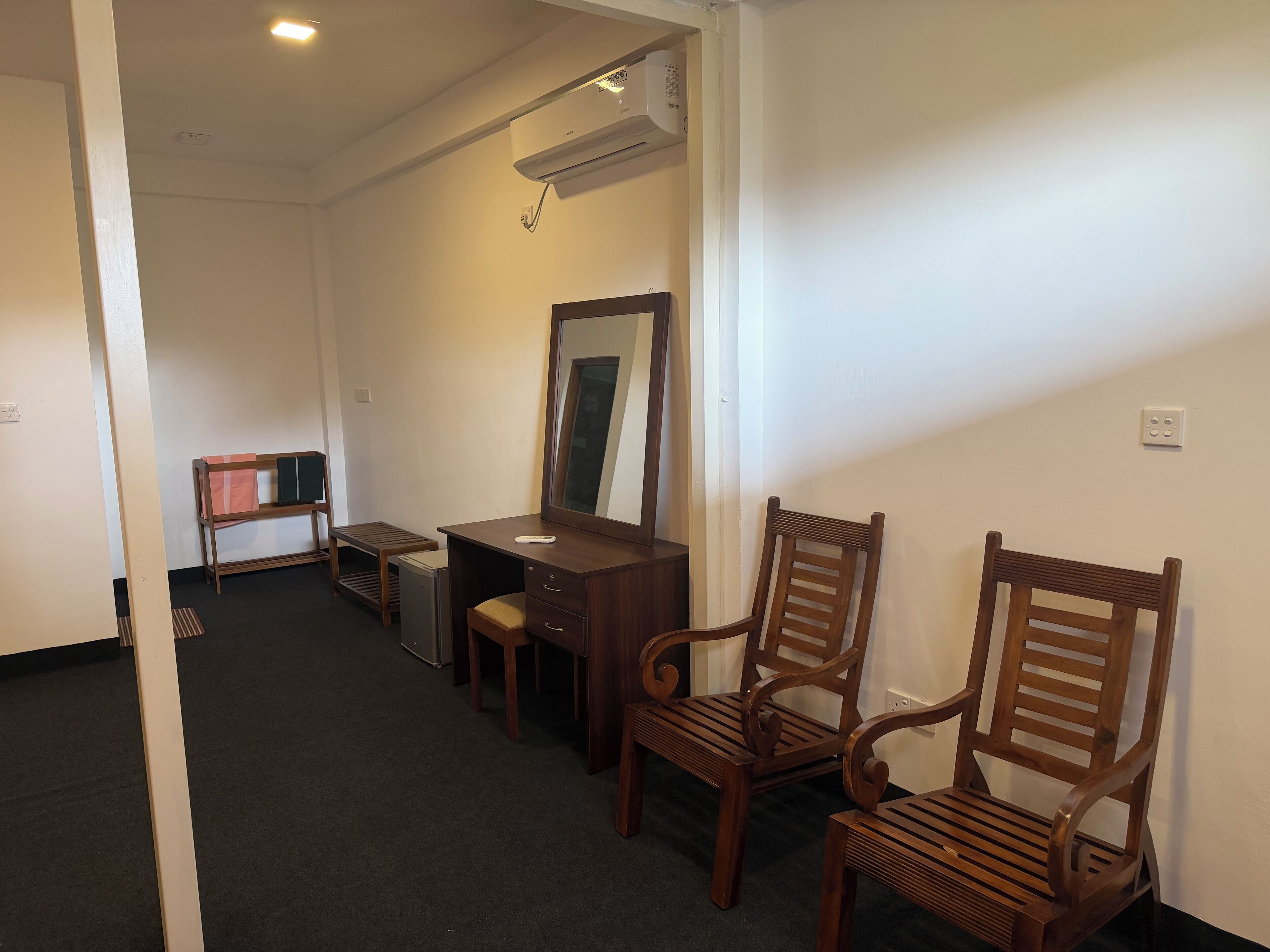 A hotel room with a desk, two wooden chairs, a small table, a mirror, and an air conditioning unit.
