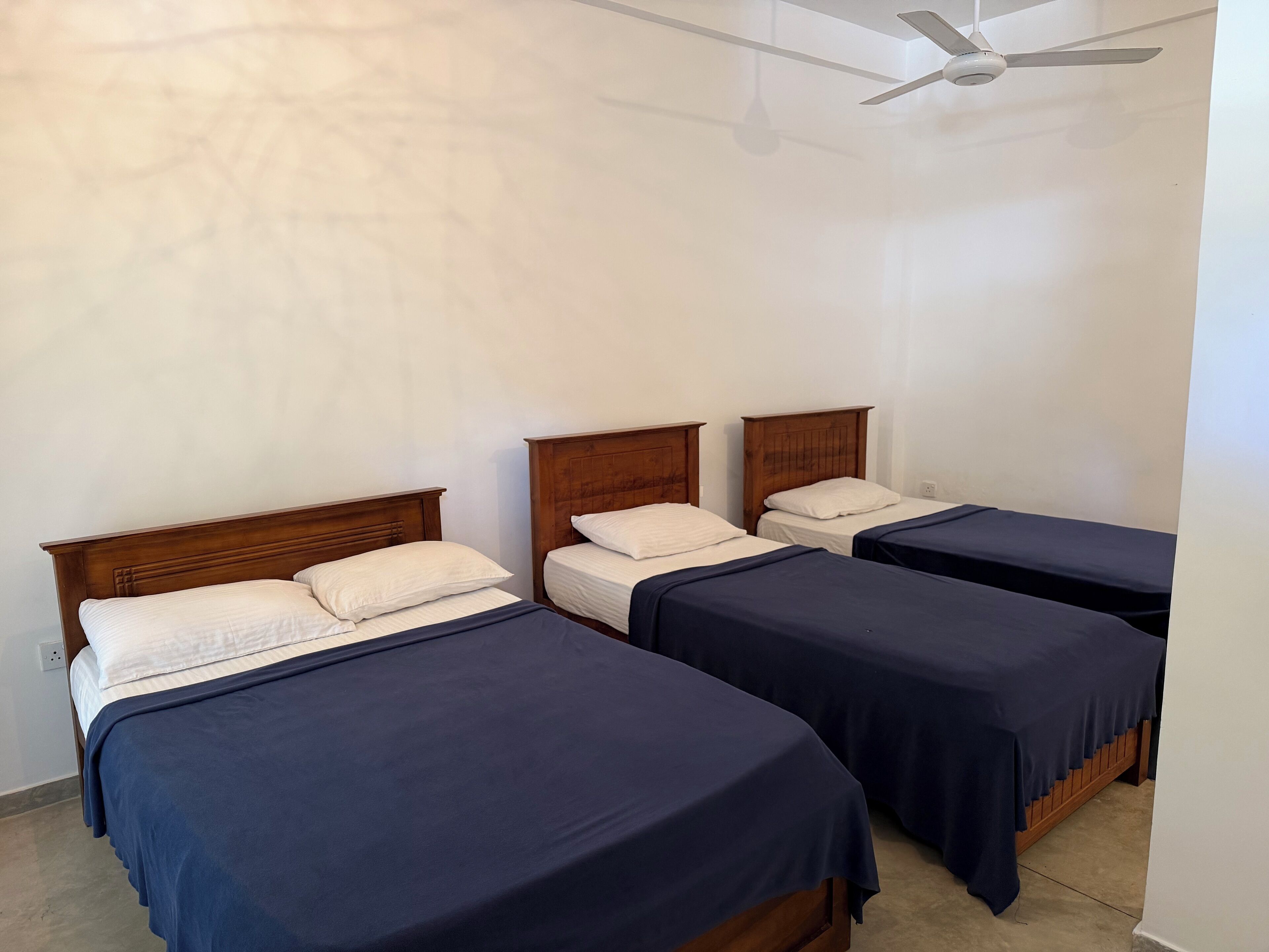 A room with three single beds, each with a wooden headboard and a dark blue bedspread. There is a ceiling fan above the beds.