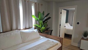 A bedroom with a wooden bed, white bedding, a large green plant, and a view into an adjacent room.