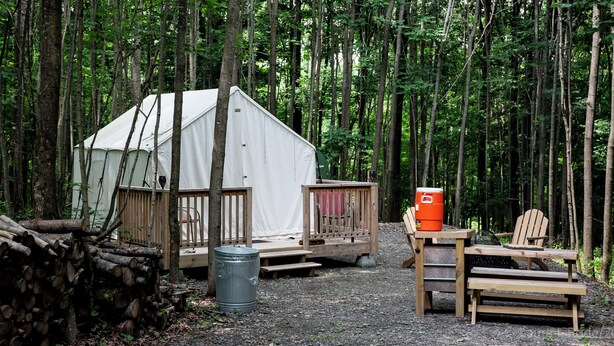 A forest campsite with a tent, a wooden deck, a metal barrel, and a table with chairs. - Charming Campsite in Hartwick, NY few miles from Cooperstown & area attractions (Hartwick)