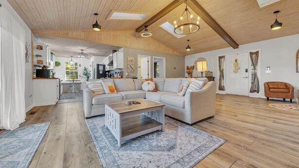 A spacious living area with a high ceiling, wooden beams, a large sofa, a coffee table, and a kitchen in the background.