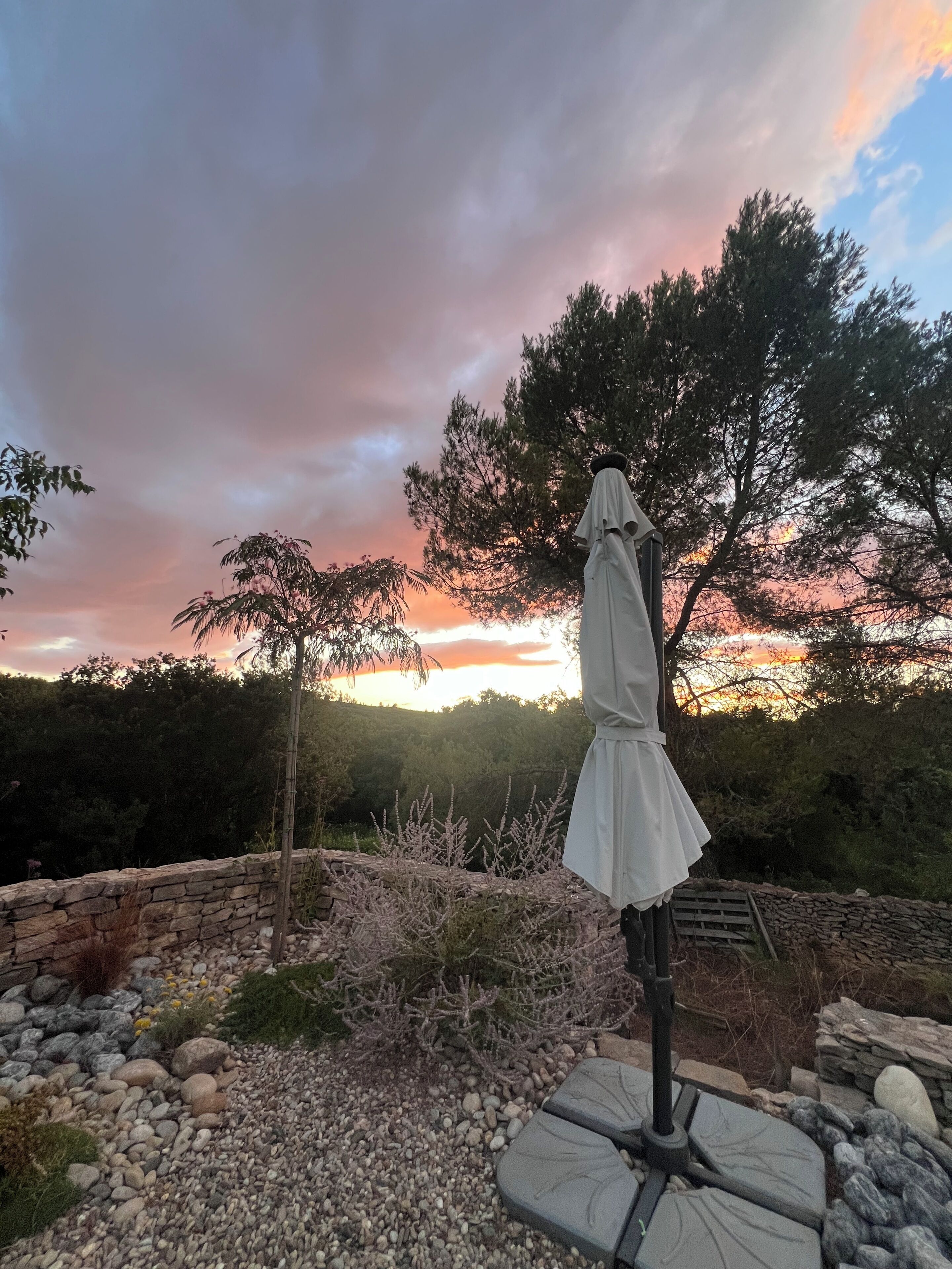A statue of a figure in a white dress, surrounded by a stone wall and gravel path, with a backdrop of trees and a sunset sky.
