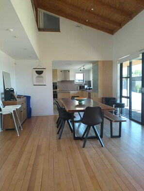 A modern kitchen with wooden cabinets, a dining area with a large table and chairs, and a view of the outdoors through large windows.