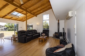 A spacious living room with a high ceiling, wooden beams, a black leather sofa, a black leather recliner, a wooden coffee table, and a black stove.