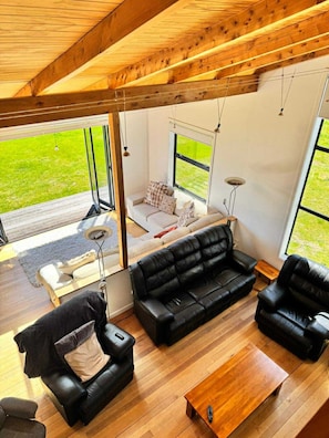 A modern living room with a wooden ceiling, black leather sofas, a wooden coffee table, and large windows offering a view of a grassy area.