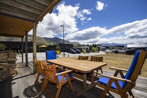 An outdoor seating area with a wooden table and chairs, a covered patio, and a view of a parking lot with vehicles and mountains in the background.