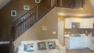 A living room with a wooden staircase, a sofa with patterned cushions, a kitchen with white cabinets, and a clock on the wall.