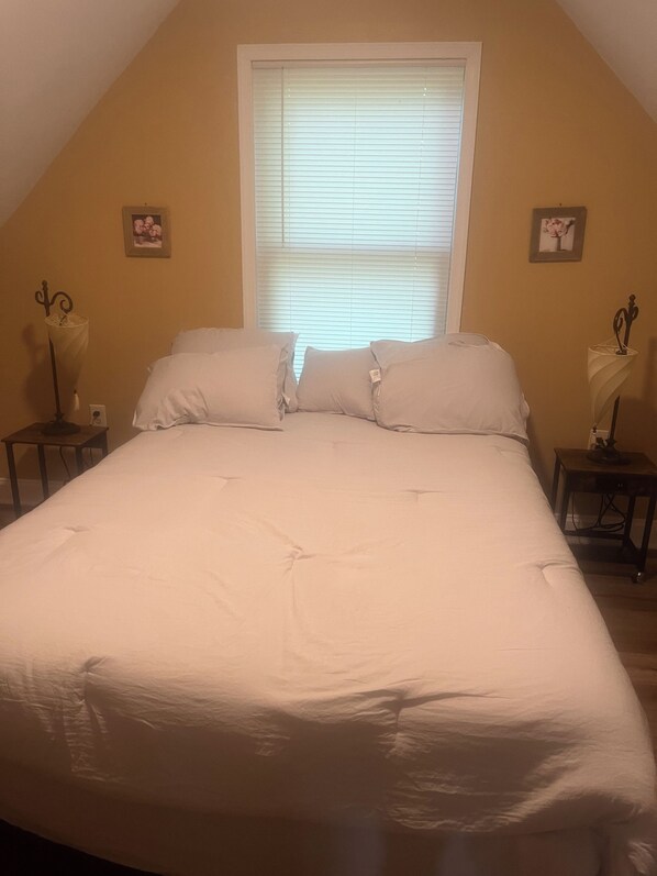 Une chambre avec un grand lit, deux tables de chevet, une fenêtre avec des stores et deux tableaux accrochés au mur.