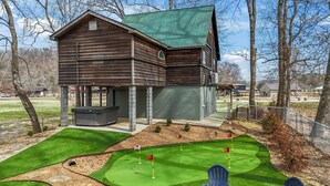 A wooden cabin with a green roof, a patio, and a small putting green with three flags.