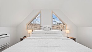 A bedroom with a high ceiling, a bed with white bedding, two bedside lamps, and a window with a view of bare trees.