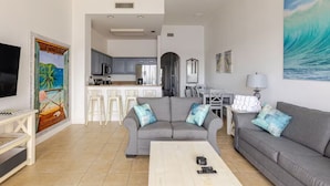 A living room with a grey sofa, a coffee table, and a TV. There is a kitchen in the background with grey cabinets and a dining area with a table and chairs.