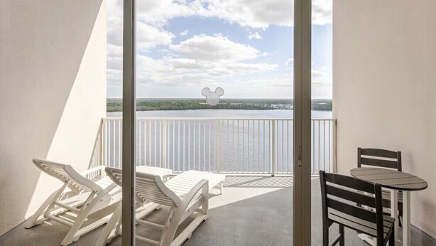 A balcony with white chairs and a table, overlooking a body of water and a distant landscape.