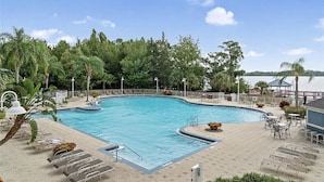 A swimming pool with a curved edge, surrounded by lounge chairs and tables, with a view of a body of water and trees in the background.