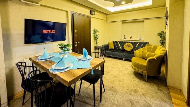 A modern room with a wooden dining table, black chairs, a sofa, and a yellow armchair. A flat-screen TV is mounted on the wall, and there is a ceiling light fixture.