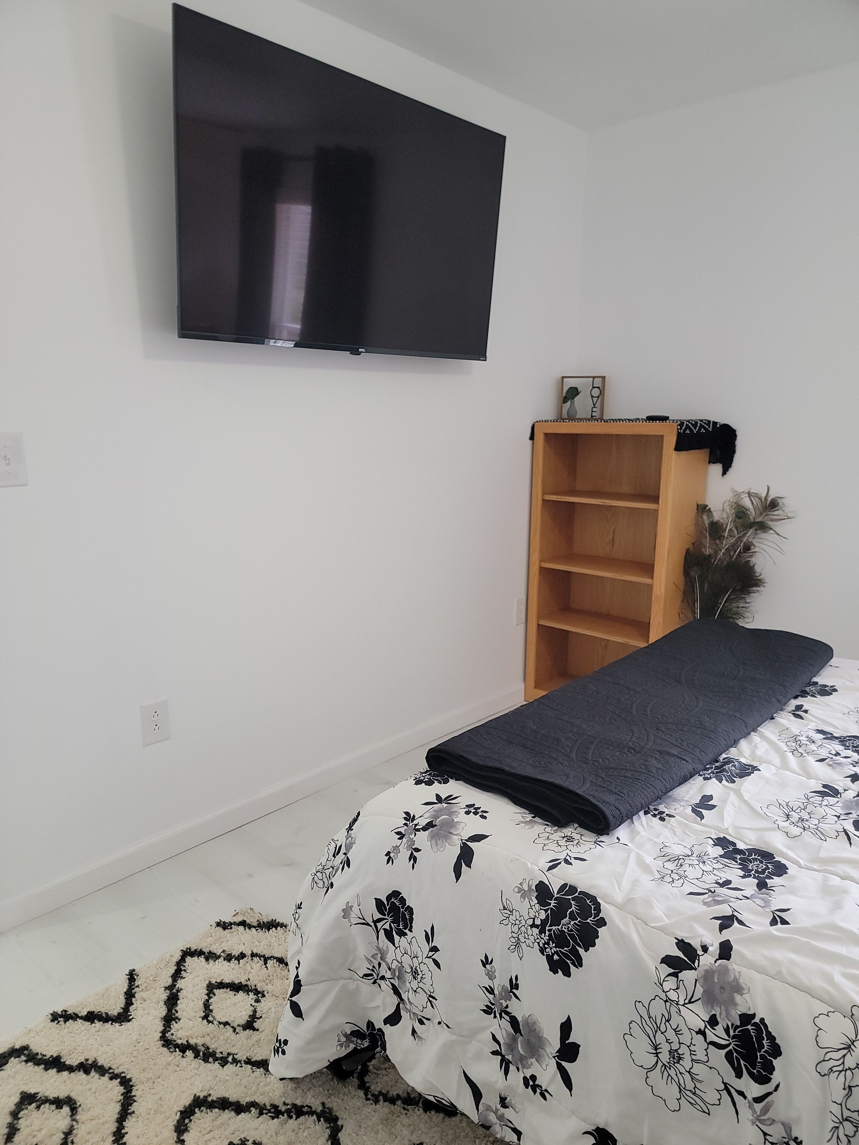 A bedroom with a bed, a TV mounted on the wall, a wooden bookshelf, and a decorative plant.