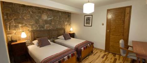Una habitación con dos camas, una pared de piedra, una puerta de madera y una pequeña mesa con una silla.