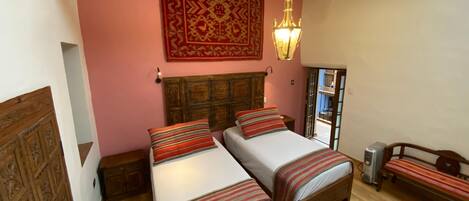 A bedroom with two beds, a wooden headboard, a red patterned tapestry, a hanging light fixture, and a wooden bench.