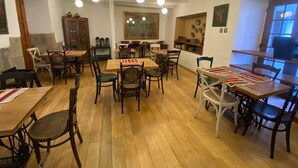 Un espace repas avec plusieurs tables et chaises en bois, un coin cuisine à l’arrière-plan et un éclairage suspendu.