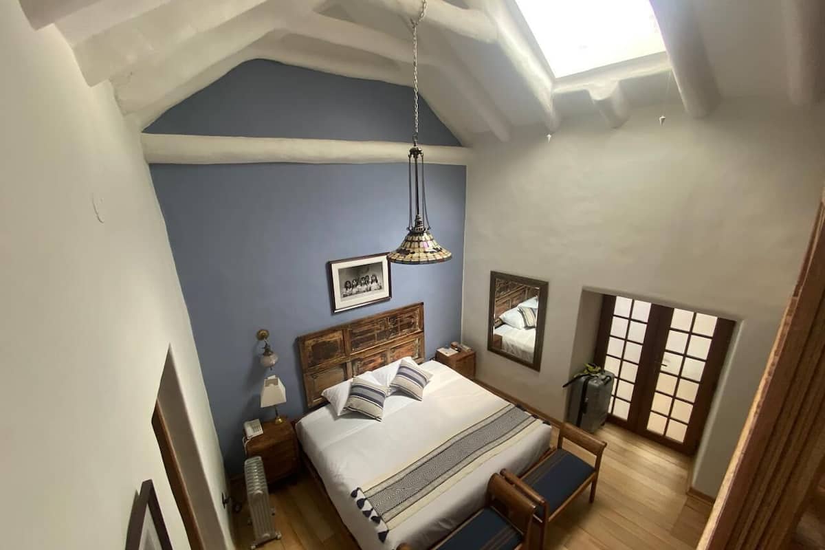 Un dormitorio con una cama grande, cabecera de madera, mesita de noche, lámpara colgante, ventana con vistas y suelo de madera.