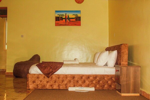 A bedroom with a bed, a bedside table, a brown ottoman, and a painting on the wall.
