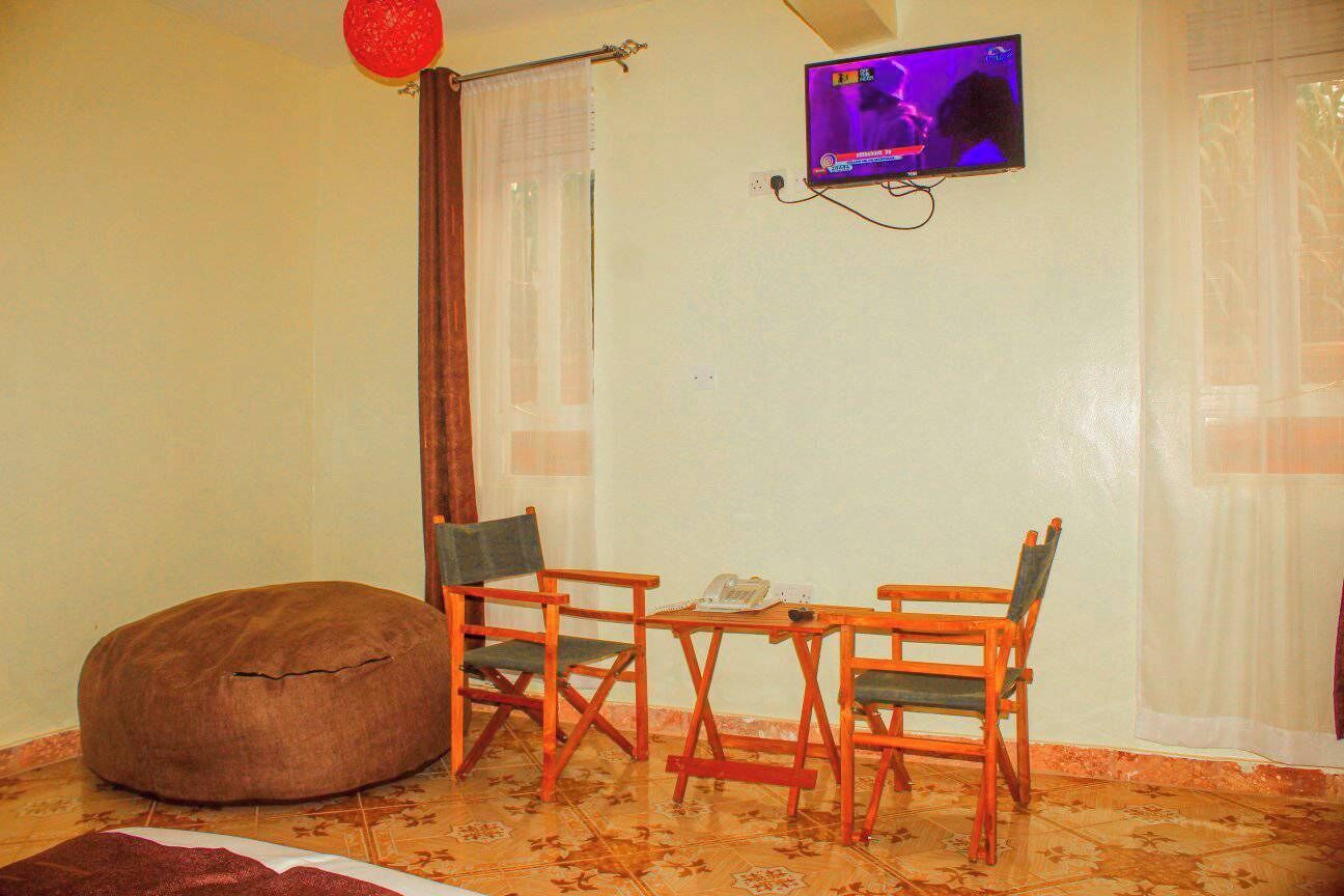 A room with a television mounted on the wall, a brown bean bag chair, two wooden chairs, a small table with a telephone, and patterned flooring.